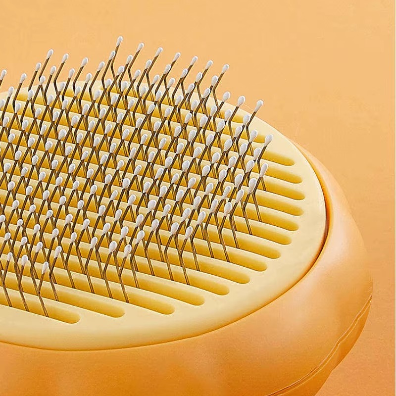 Close-up of a pet grooming brush with a pale yellow bristle pad, silver metal pins, and a peach-colored body on a matching peach background.