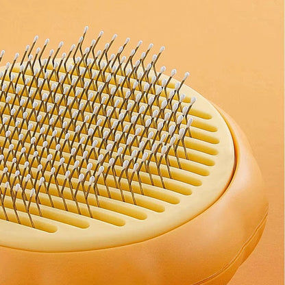 Close-up of a pet grooming brush with a pale yellow bristle pad, silver metal pins, and a peach-colored body on a matching peach background.