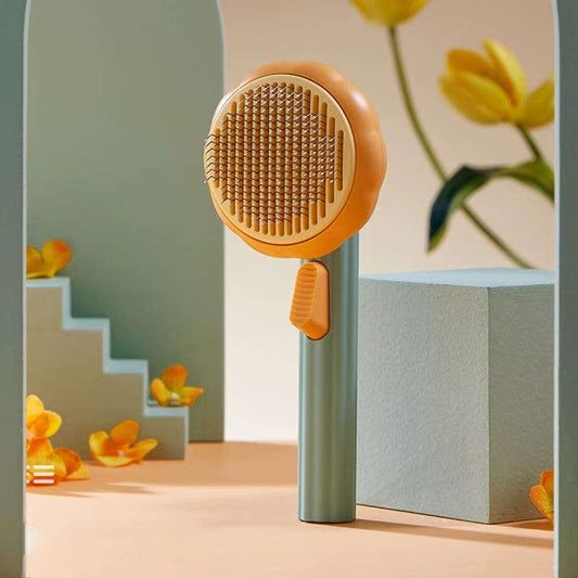 A pet grooming brush with a round orange and beige bristle head and a sage green handle, standing upright on a peach-colored surface with decorative yellow flowers and geometric props in the background.