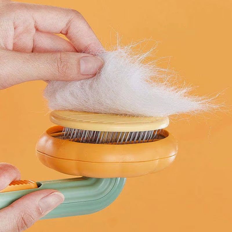 A pet grooming brush with a mint green handle, orange base, and yellow bristle top, shown with a hand pulling white fur off a dog or cat. The background is a solid orange color.